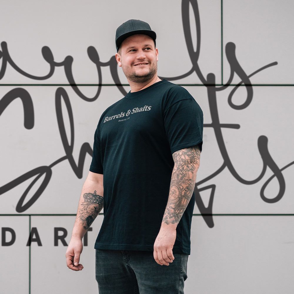 Tričko Barrels and Shafts - Černé Ein Mann trägt ein schwarzes T-Shirt mit der Aufschrift „Barrels & Shafts“. Er steht vor einer Wand mit passender Schrift im Hintergrund.