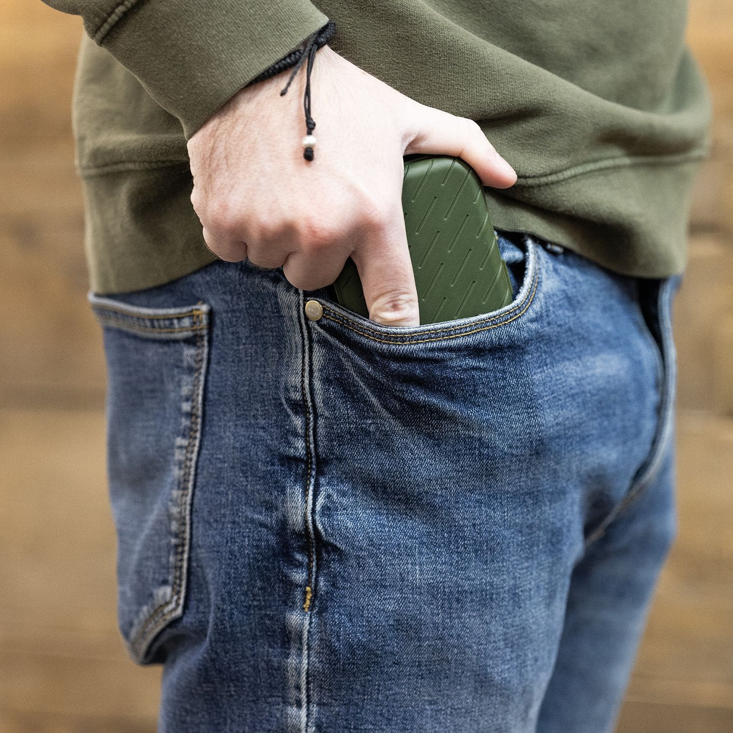 Pouzdro na šipky Harrows Slimcase Eine Person steckt ein grünes Etui in die vordere Hosentasche einer Jeans. Das Etui hat eine strukturierte Oberfläche und passt gut in die Tasche.