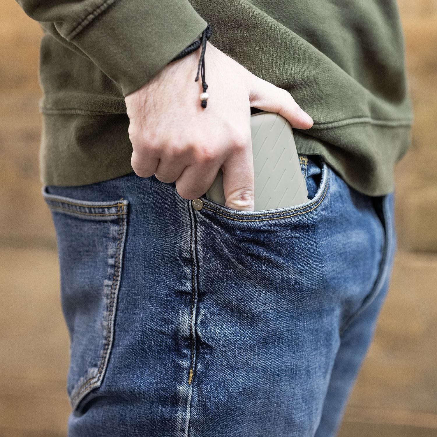 Pouzdro na šipky Harrows Slimcase Eine Person steckt eine graue Darttasche in ihre Hosentasche. Die Person trägt eine blaue Jeans und einen olivgrünen Pullover.