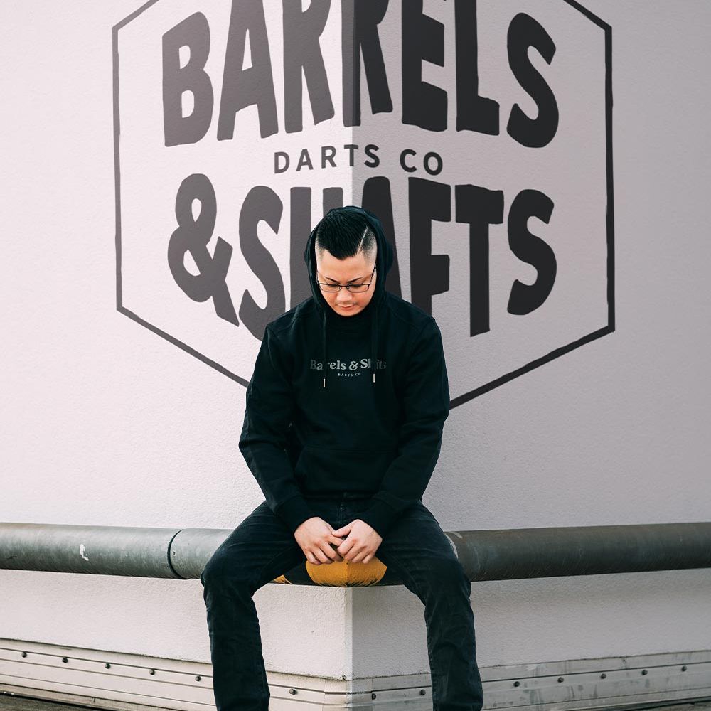 Barrels and Shafts Mikina - černá Eine Person trägt einen schwarzen "Barrels and Shafts" Hoodie und sitzt auf einer Bank. Im Hintergrund ist ein großes Wandlogo mit der Aufschrift "Barrels & Shafts Darts Co" zu sehen.