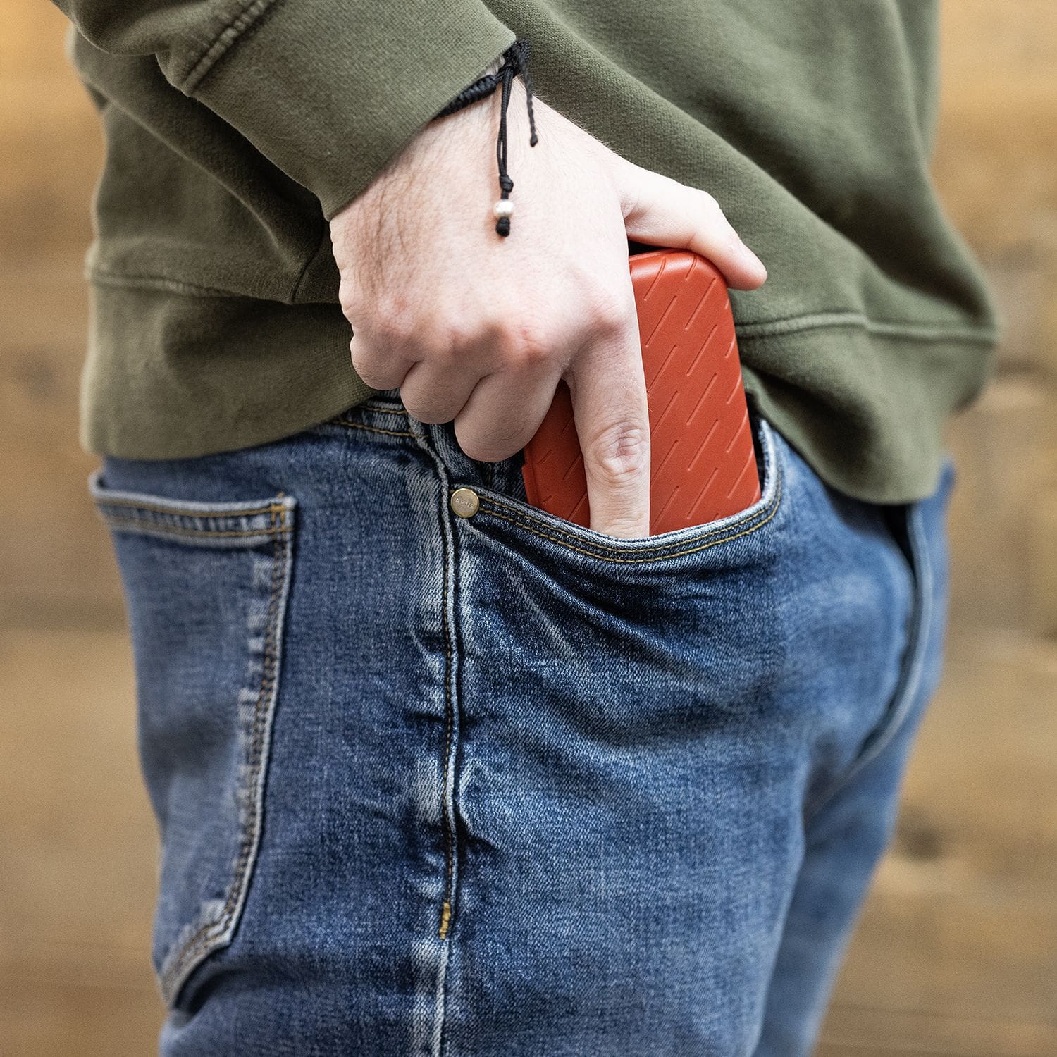 Pouzdro na šipky Harrows Slimcase Eine Person steckt ein rotes Etui in die Hosentasche. Sie trägt eine blaue Jeans und einen grünen Pullover.