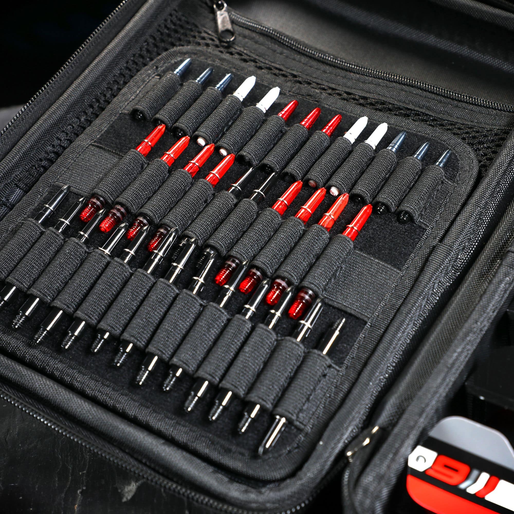 Winmau Armor G4 Dartwallet Das Bild zeigt ein geöffnetes schwarzes Dartwallet mit vielen ordentlich einsortierten Dartspitzen. Die Dartspitzen sind in elastischen Schlaufen gehalten und teilweise rot, weiß oder schwarz gefärbt.