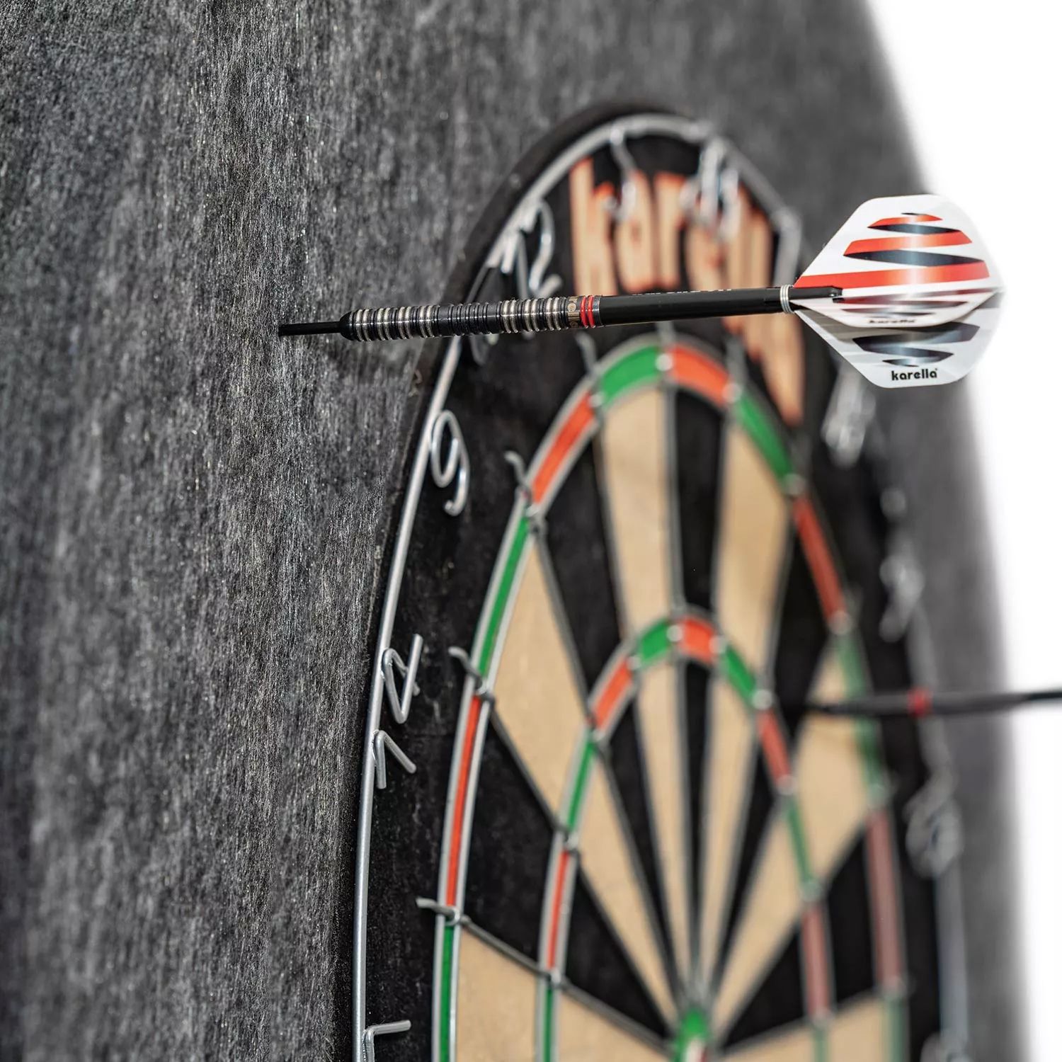 Das Bild zeigt eine Karella Ultimo Steeldartboard Surround, die einen Dartpfeil auffängt. Die graue Umrandung schützt die Wand vor Fehltreffern neben der Dartscheibe.