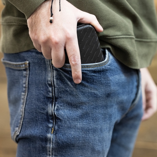 Pouzdro na šipky Harrows Slimcase Eine Person steckt ein schwarzes Etui in die Hosentasche. Sie trägt eine grüne Jacke und eine blaue Jeans.