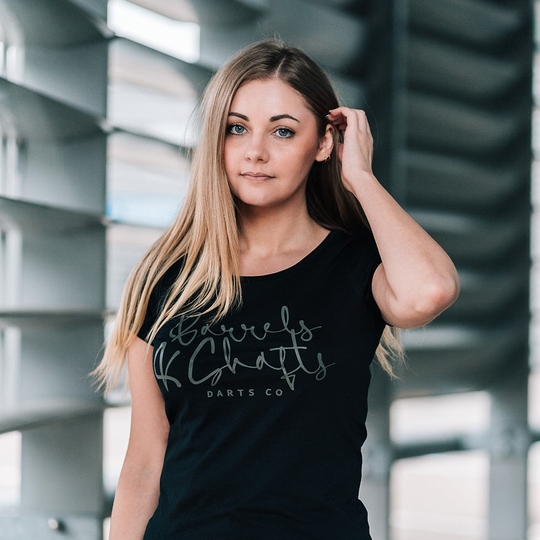 Dámské triko Barrels and Shafts – černé Eine Frau trägt ein schwarzes T-Shirt mit der Aufschrift „Barrels and Shafts Darts Co“. Sie steht vor einem modernen, lichtdurchfluteten Hintergrund.