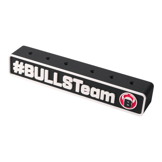 Das Bild zeigt einen schwarzen Darts-Halter mit der Aufschrift "#BULLSTeam" und einem roten Bullen-Logo. Der Halter hat mehrere Löcher zur Aufbewahrung von Dartpfeilen.