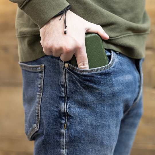 Pouzdro na šipky Harrows Slimcase Eine Person steckt ein grünes Etui in die vordere Hosentasche einer Jeans. Das Etui hat eine strukturierte Oberfläche und passt gut in die Tasche.