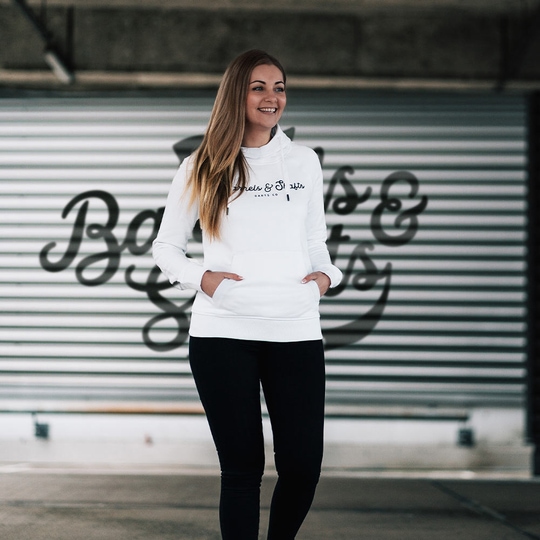 Dámská mikina s kapucí Barrels and Shafts – bílá Eine Frau trägt einen weißen "Barrels and Shafts" Damen Hoodie. Sie steht vor einem Hintergrund mit großem Schriftzug und lächelt.